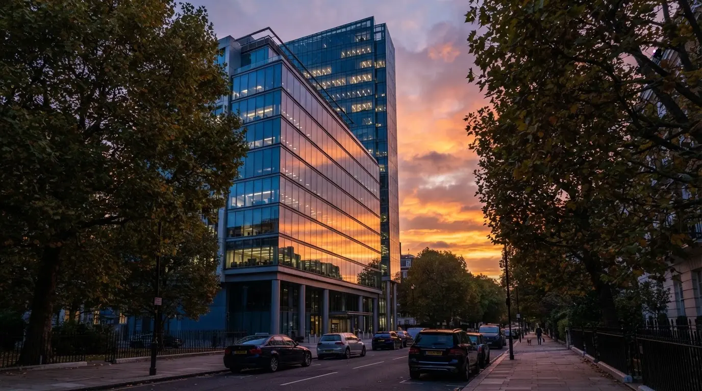 Edifício de escritórios com fachada de vidro ao entardecer, reflexo do céu cor-de-laranja e uma rua tranquila em primeiro plano.